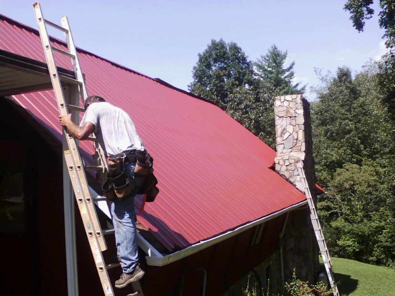 Roofer on a ladder beside a red metal roof and stone chimney — Soffit & Fascia Repair by Roofing Brothers in Crossville